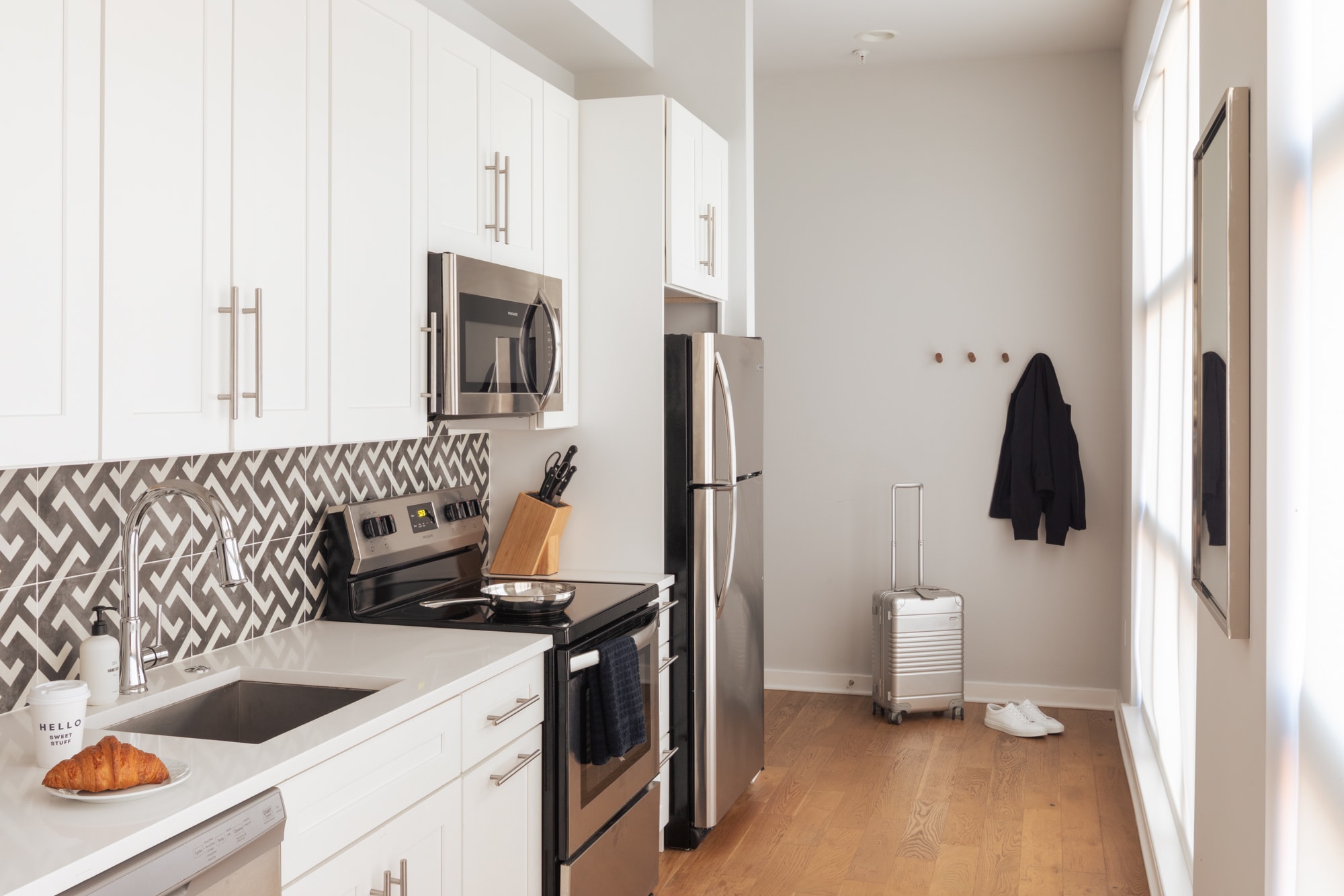Contemporary kitchen with white shaker cabinets, stainless steel appliances including a microwave, stove, and refrigerator, and a gray and white herringbone backsplash. A sink with a gooseneck faucet is visible, and a croissant sits on the counter.