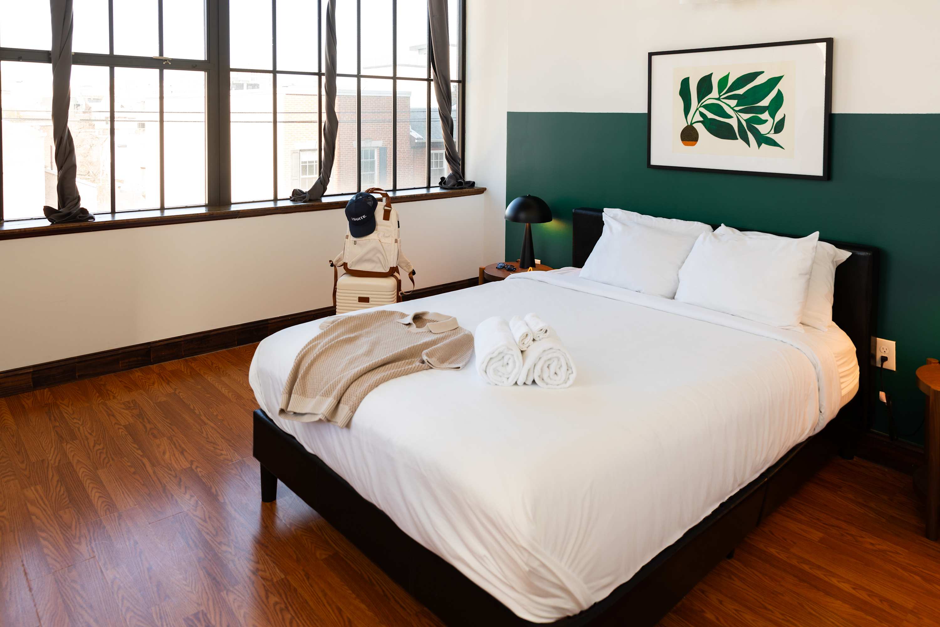 Cozy bedroom with a queen-sized bed dressed in white linens and a beige throw, flanked by a dark nightstand with a lamp. Large industrial windows with dark curtains frame the view, complementing the two-toned white and dark green walls and warm hardwood floors.