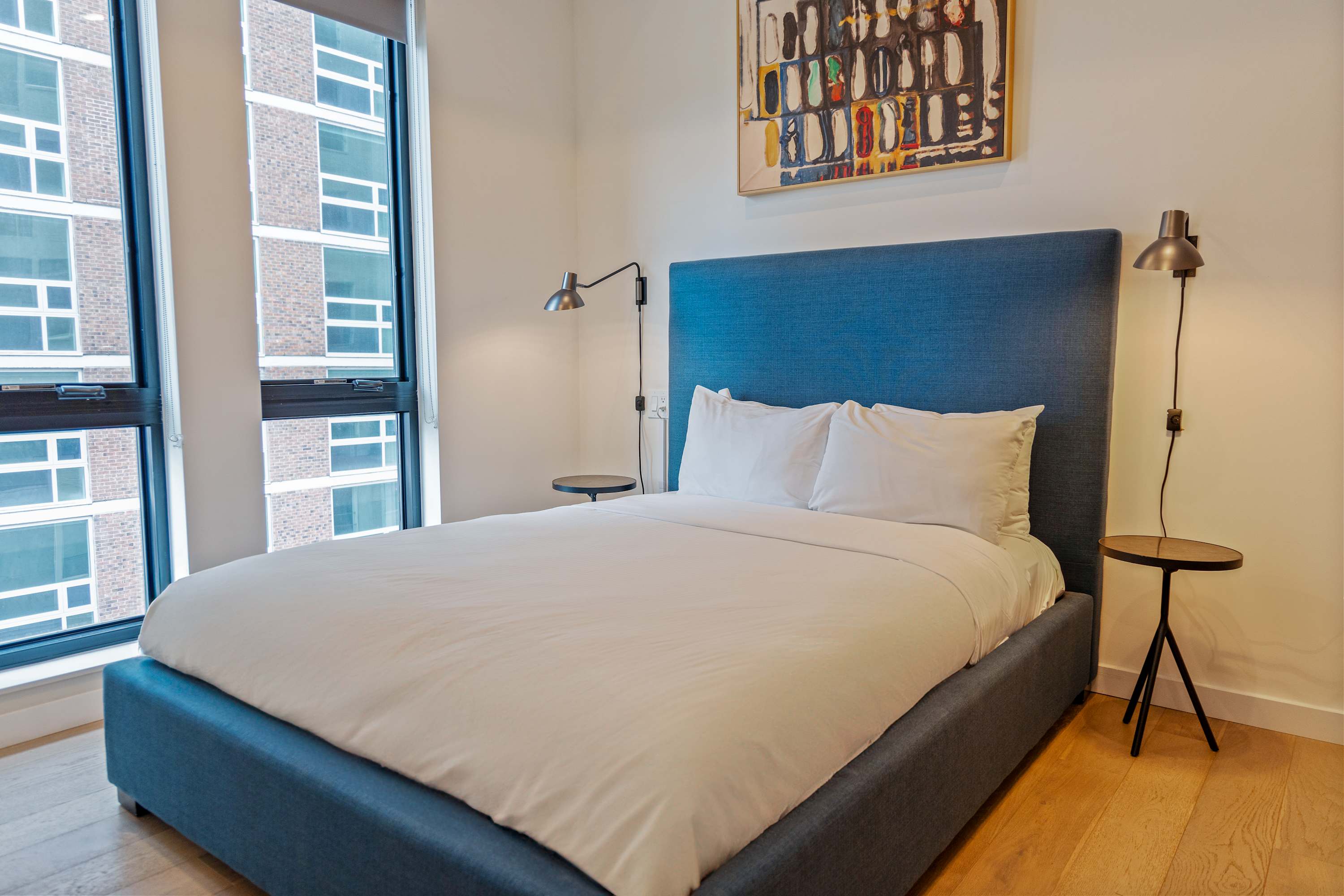 Bright bedroom featuring a queen-sized bed with a blue upholstered headboard and crisp white bedding, flanked by two small round side tables with wall-mounted lamps. Large windows with dark blinds offer a view of an adjacent building.