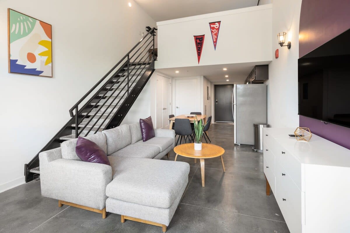 A view of the open-concept living space with a grey sectional sofa, a round wooden coffee table, and a dining area with a wooden table and dark chairs. A black metal staircase leads to a loft area.