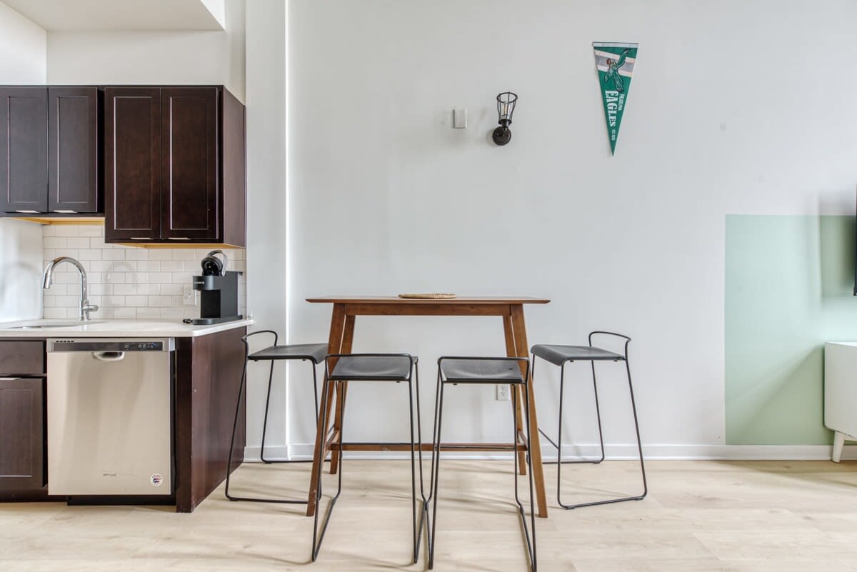 Compact kitchen area with dark wood cabinets, white subway tile backsplash, and stainless steel appliances including a dishwasher and sink. A wooden high-top dining table with four black bar stools is adjacent to the kitchen.