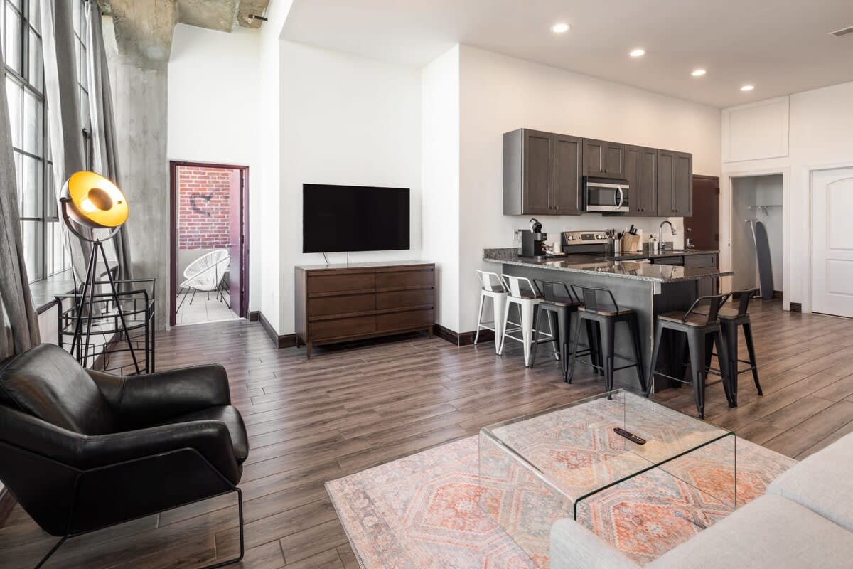 Spacious open-plan living room with a black leather armchair, a large wall-mounted TV above a wooden dresser, and a view into the kitchen area with bar seating. A balcony is visible through an open doorway.