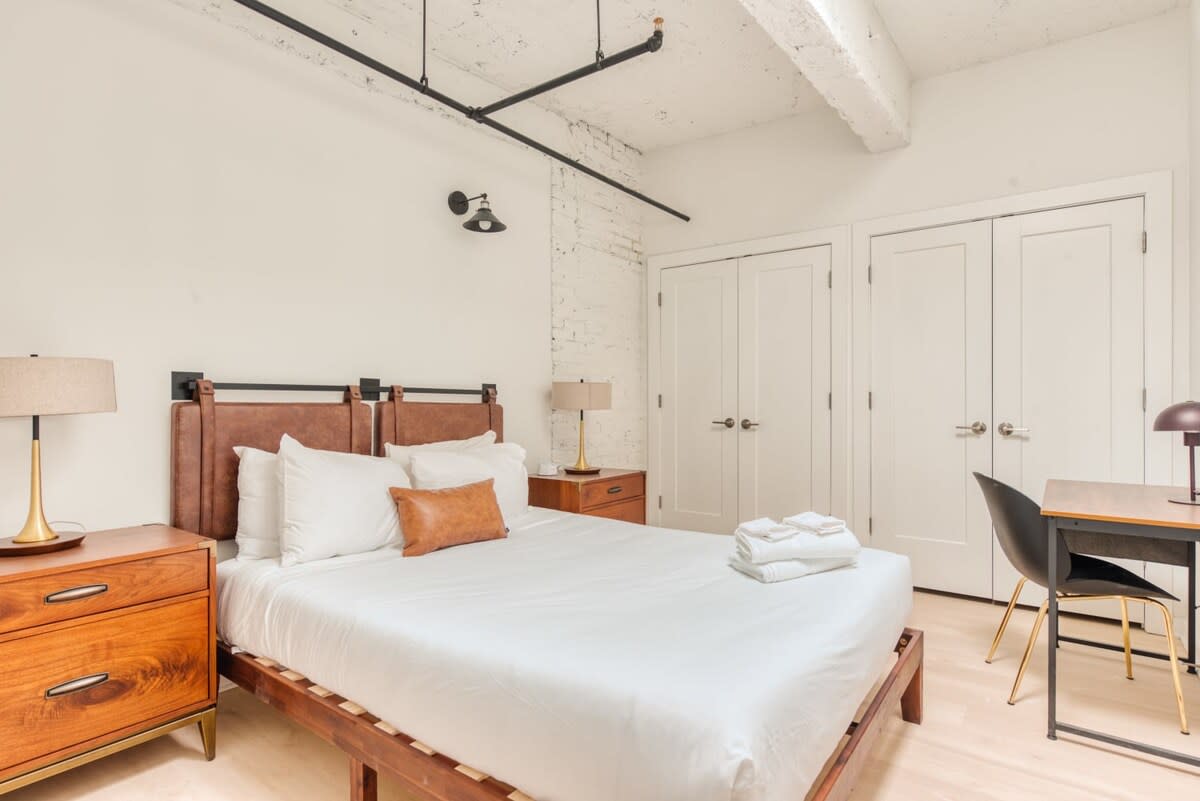 Bedroom with a queen bed featuring a brown leather headboard, flanked by wooden nightstands with lamps, a small desk and chair, and white double closet doors, under an exposed ceiling with pipes.