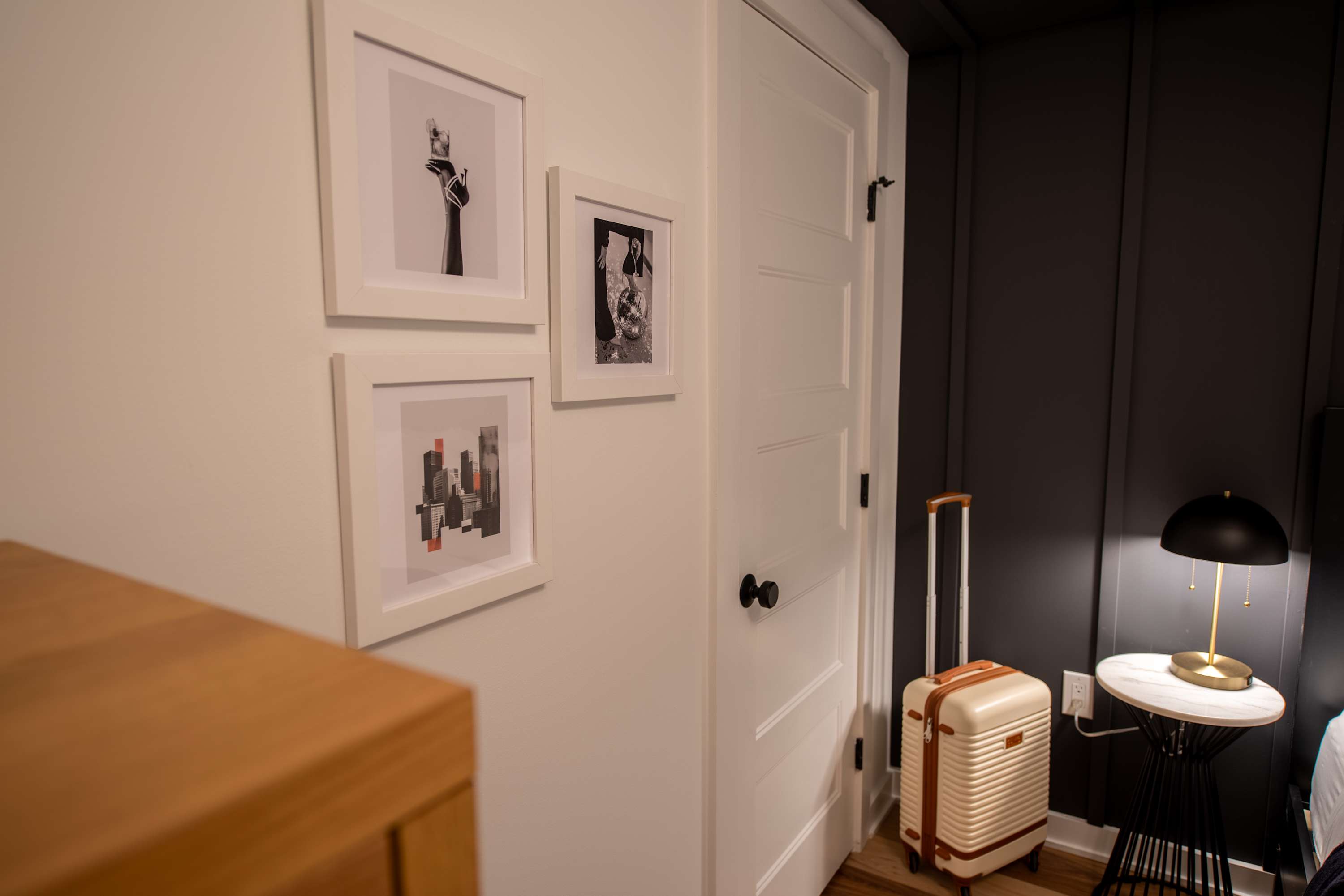 A corner view of a modern bedroom with a white door, dark paneled accent wall, and a nightstand with a lamp, featuring framed artwork on the white wall and a suitcase on the floor.