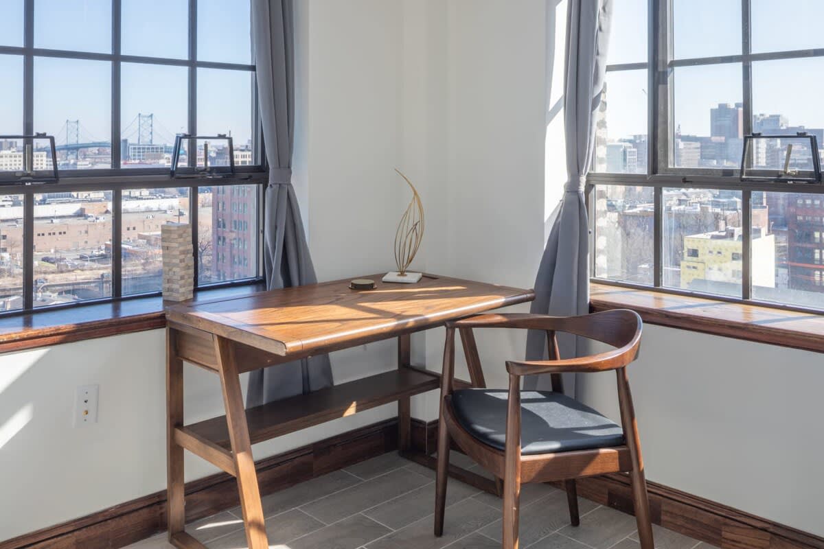 A minimalist wooden desk and matching chair are positioned in a corner with large grid windows, offering panoramic city views including a distant bridge. A decorative sculpture adorns the desk, bathed in natural light.