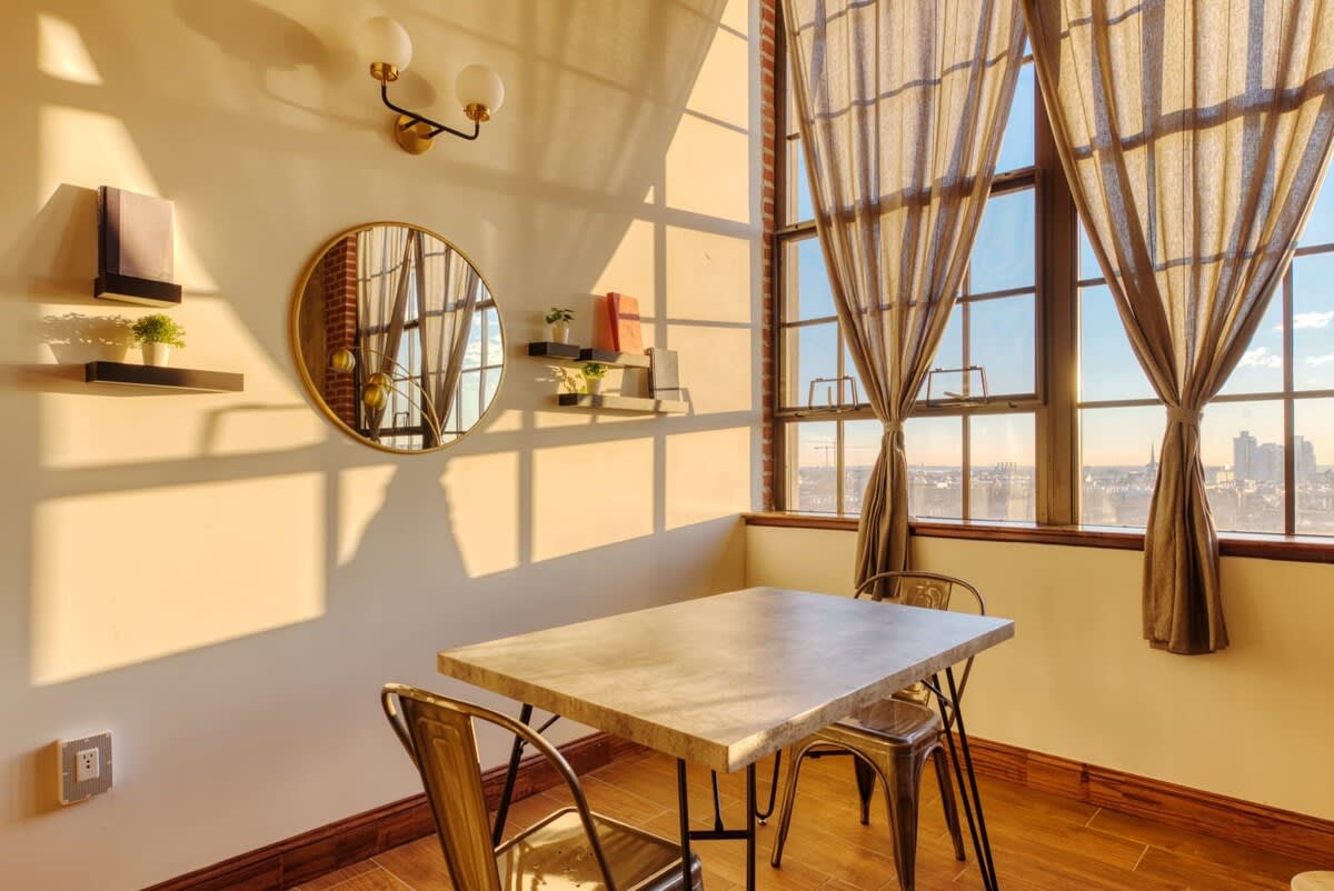 A cozy dining nook with a rectangular table and two industrial-style metal chairs, positioned by large windows with city views. The wall is decorated with a round gold-framed mirror and floating shelves displaying small plants and books.