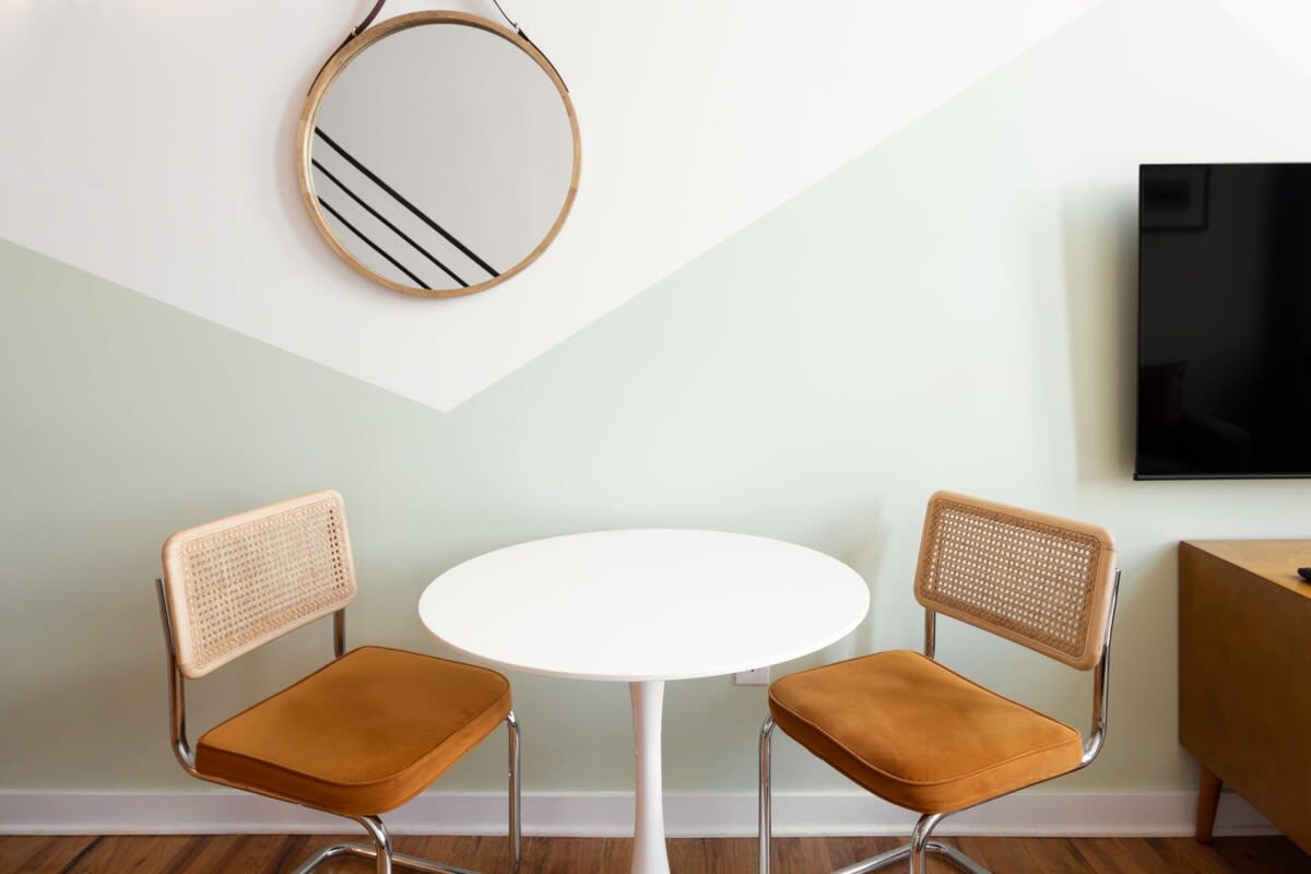A small, modern dining area featuring a round white table and two cane-back chairs with brown cushions. A round wooden mirror with a leather strap hangs on the wall, which has a geometric light green and white paint design.