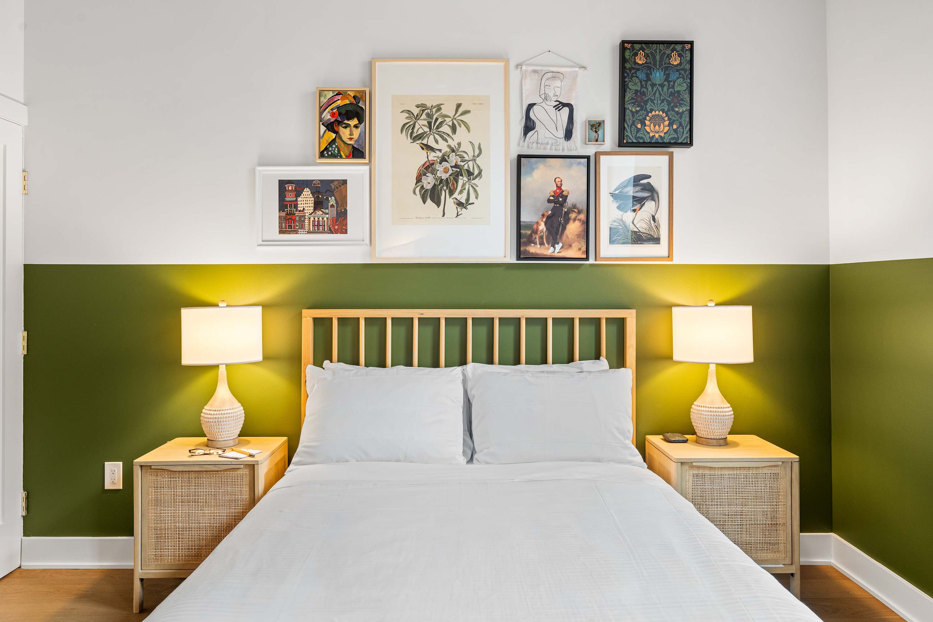 A cozy bedroom featuring a queen-sized bed with white linens and a light wood slatted headboard. Two woven nightstands with white ceramic lamps flank the bed, beneath a vibrant gallery wall on a two-tone green and white wall.