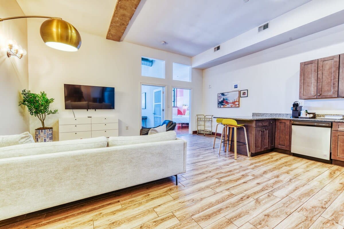 An open-concept living space with light wood-look flooring, featuring a light grey sofa and a dark leather armchair on a patterned rug, facing a wall-mounted TV above a white dresser. A modern kitchen with dark wood cabinets, granite countertops, and stainless steel appliances is visible to the right, with a breakfast bar and yellow stools.