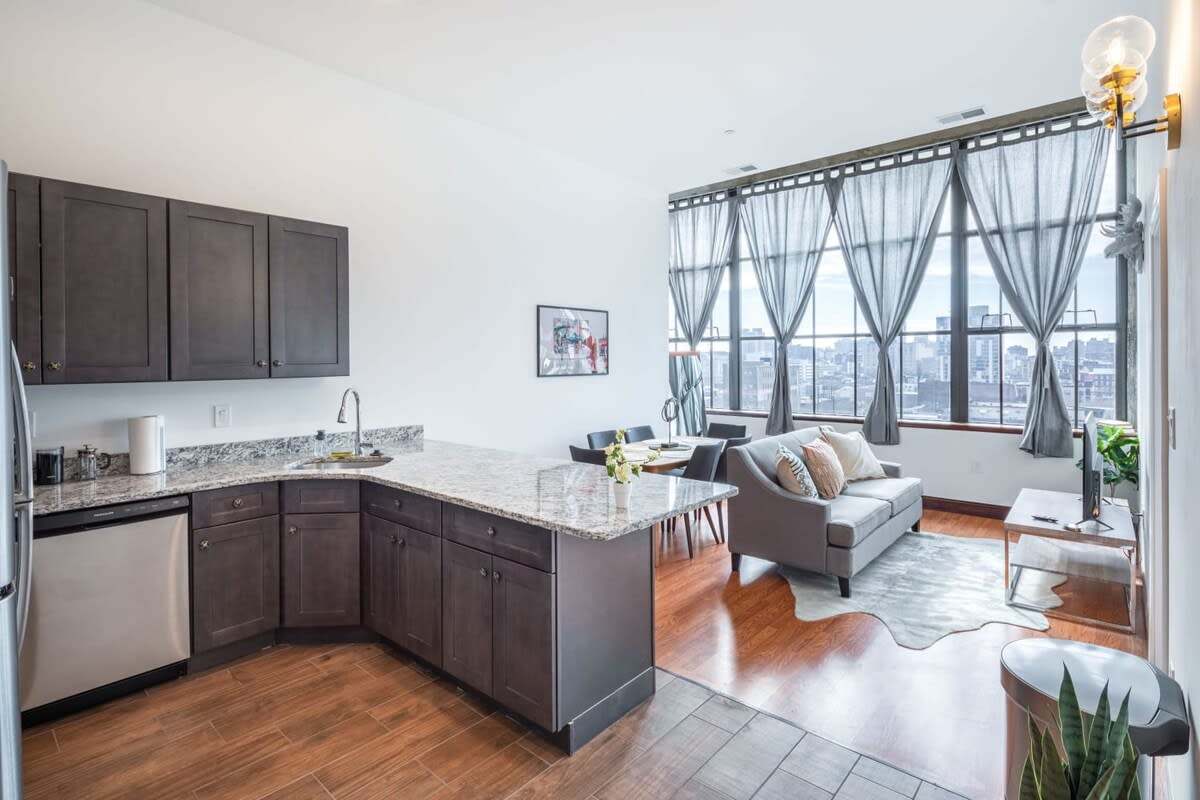Wide view of the open-concept living space, seamlessly connecting the modern kitchen with dark cabinets and granite countertops, the dining area, and the living room with a grey sofa and expansive city views through large windows.