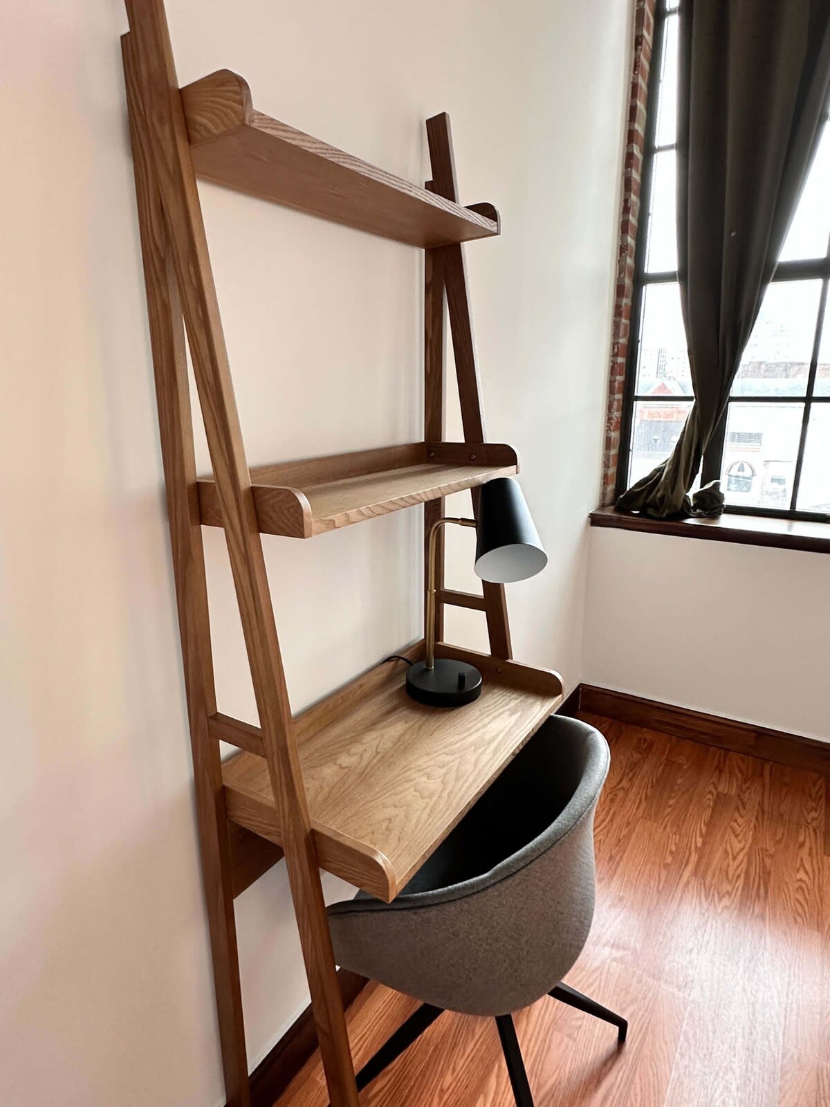 A minimalist wooden ladder desk with shelves and a black desk lamp, paired with a grey upholstered chair. A large window with dark curtains is visible to the right, offering natural light.