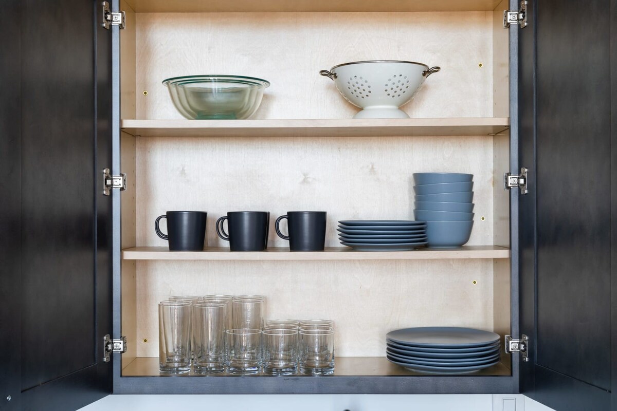 Interior view of an open kitchen cabinet, revealing neatly organized shelves stocked with a variety of dishware including clear glass bowls, a white colander, black mugs, stacks of grey plates, and several drinking glasses.
