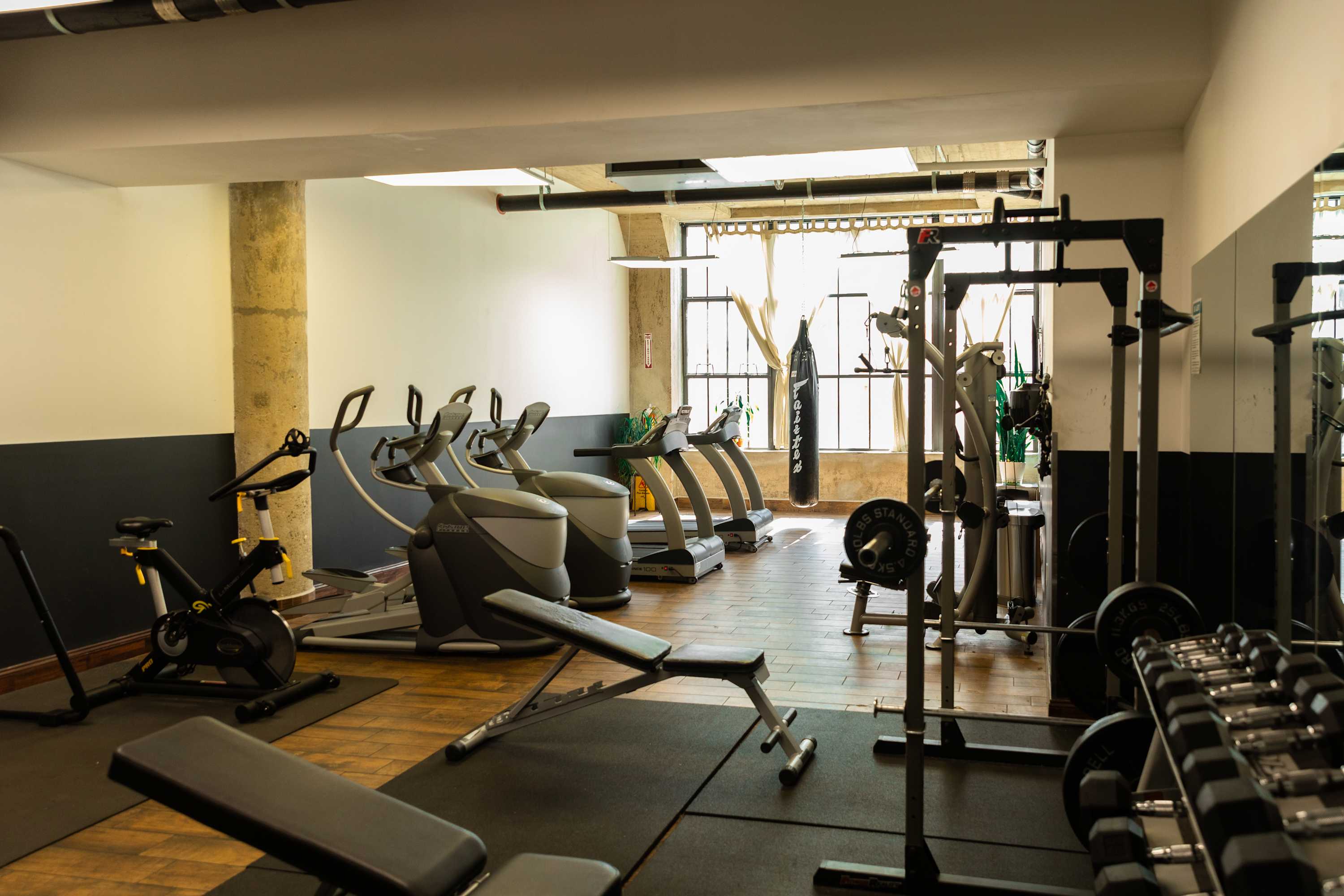 A well-equipped gym with various exercise machines including treadmills, ellipticals, and a weight bench with dumbbells, set on a dark rubber floor with large windows providing natural light.