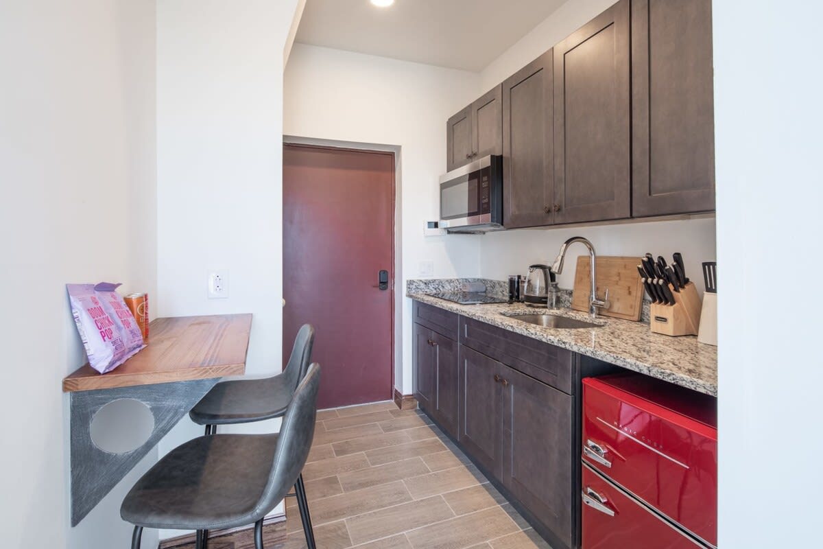 View of the kitchen area featuring dark wood cabinets, granite countertops, and a built-in bar counter with two black bar stools. A red mini-fridge is visible, and a microwave is mounted above the stove, leading to a doorway.