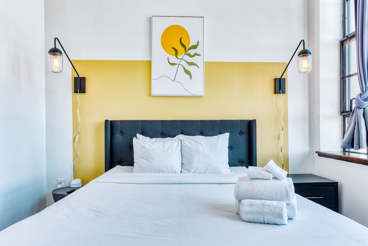 Close-up of a queen bed with a dark upholstered headboard, crisp white bedding, and a stack of fresh white towels. A yellow accent wall with modern abstract art and two black wall sconces provides a stylish backdrop.