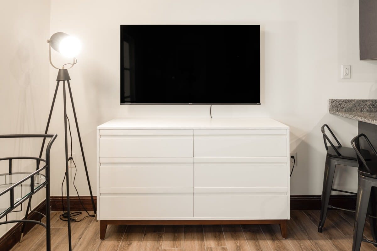 A detailed view of the living room's entertainment setup, showcasing a large wall-mounted flat-screen television positioned above a modern white six-drawer dresser with wooden legs. A stylish black tripod floor lamp stands to the left, providing ambient lighting to the space.
