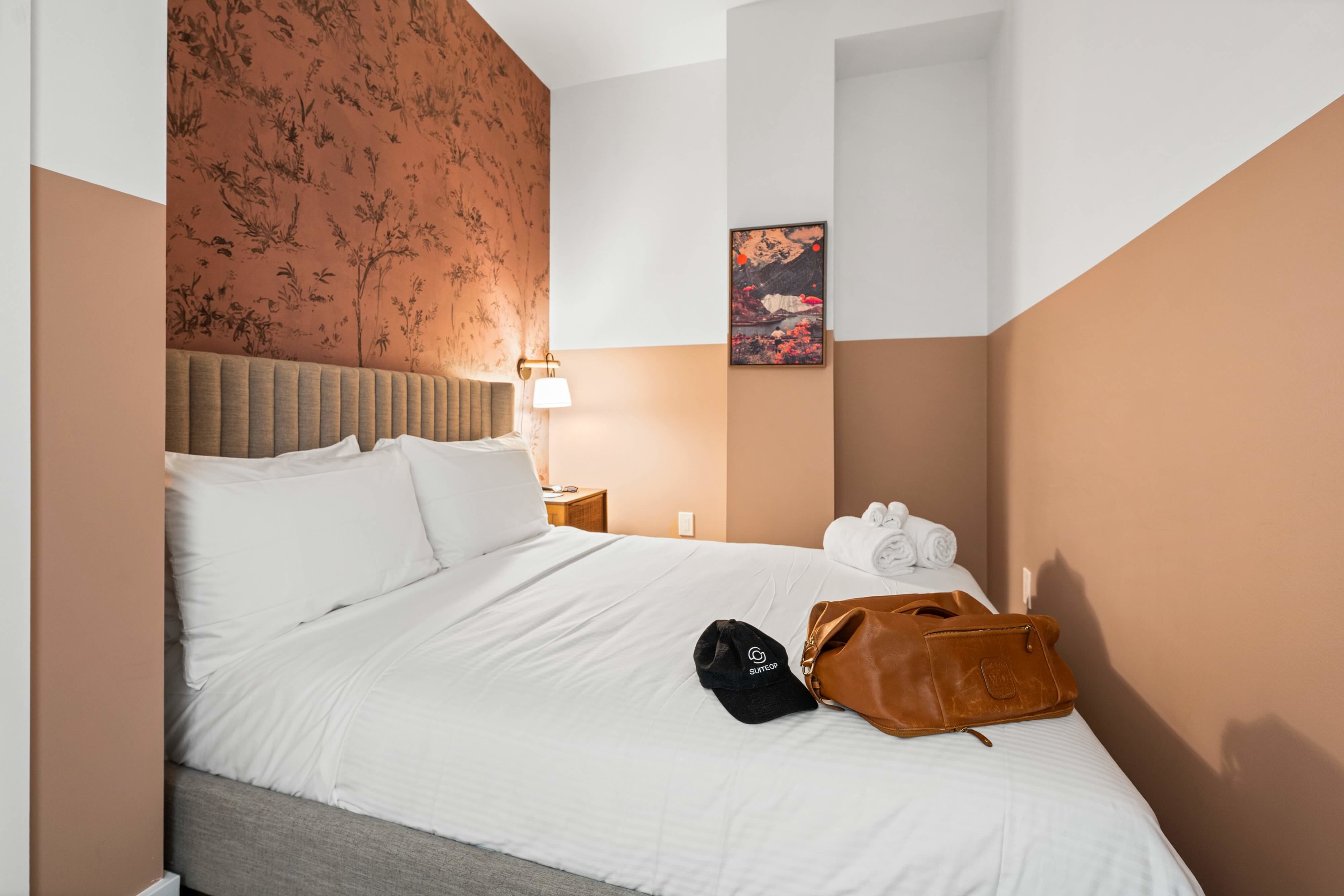 Second bedroom with a queen bed featuring a grey fabric headboard and white bedding. The walls are painted in a two-tone design with white and terracotta, and a feature wall with botanical patterned wallpaper.