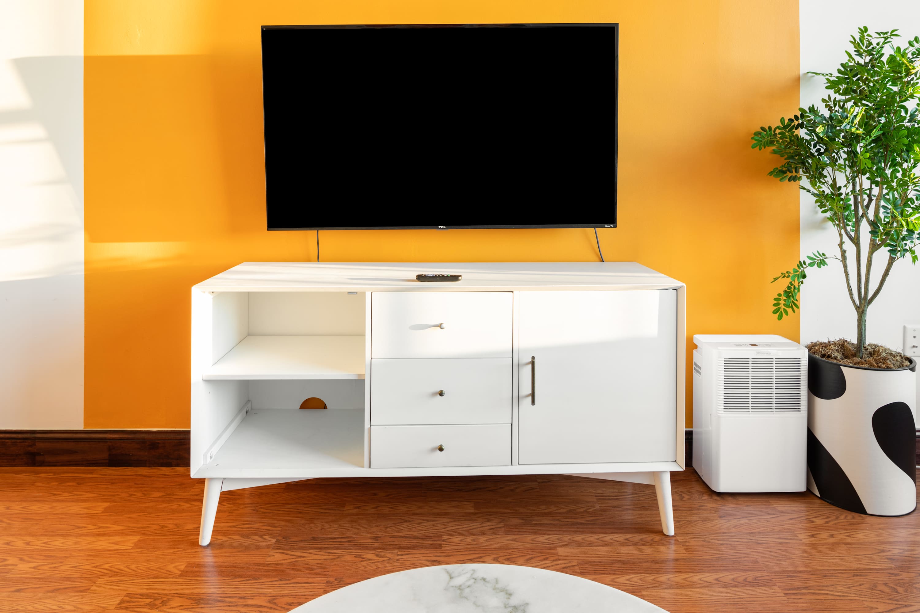 Modern white dresser with multiple drawers and shelves, supporting a wall-mounted flat-screen TV, set against a vibrant orange accent wall. A potted plant and a portable air conditioner are visible beside it.