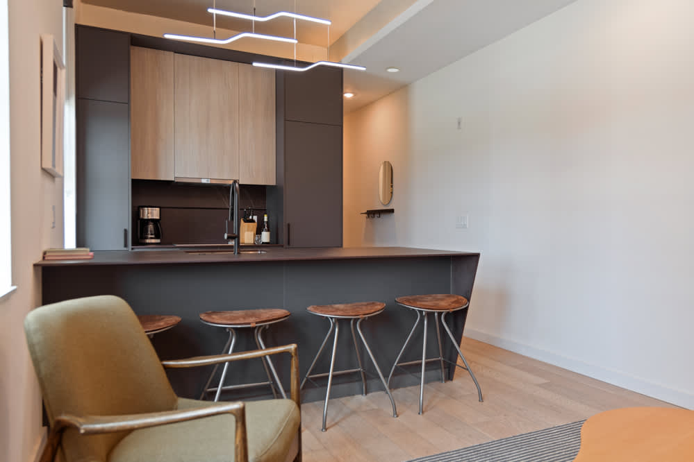 A sleek, modern kitchen with dark lower cabinets and light wood upper cabinets, featuring a large dark island with four bar stools. The open space flows into a living area with an olive green armchair and a light wood floor.