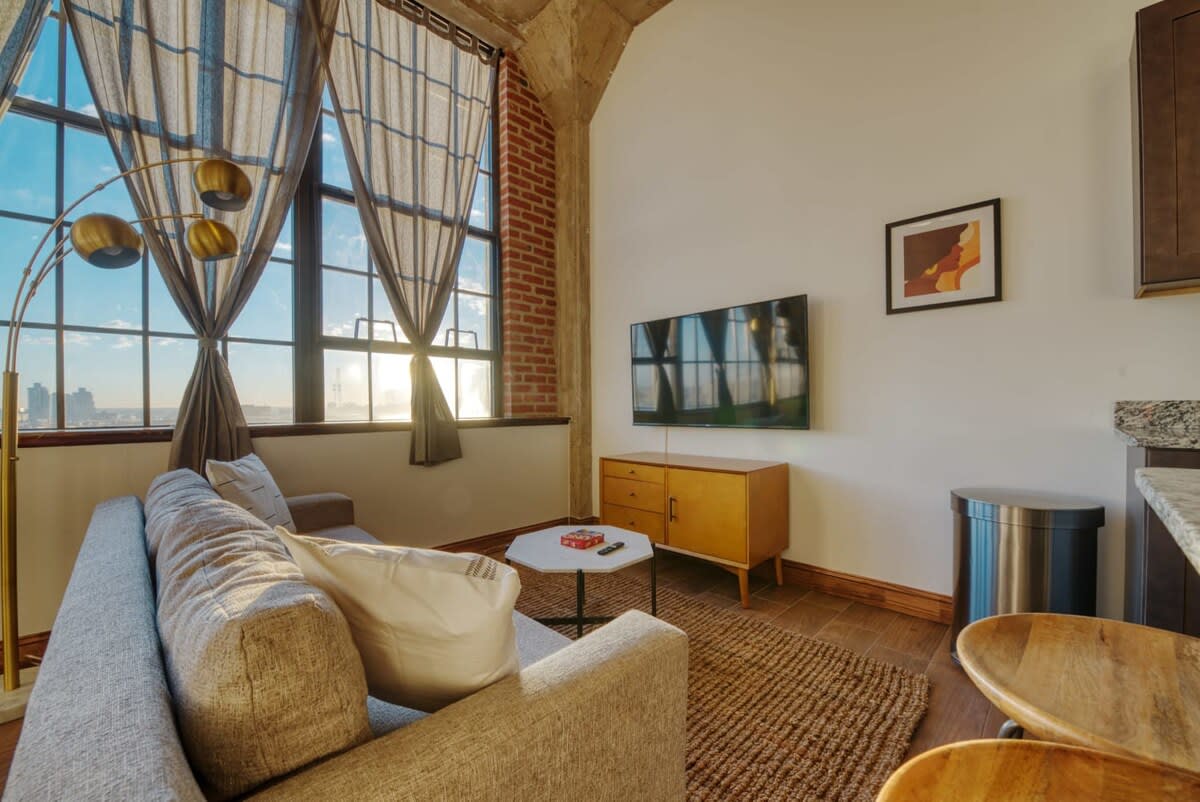 A view of the living room showcasing a wall-mounted TV above a mid-century modern wooden console. A light gray sofa and a hexagonal coffee table define the seating area, with large windows providing ample natural light and city views.