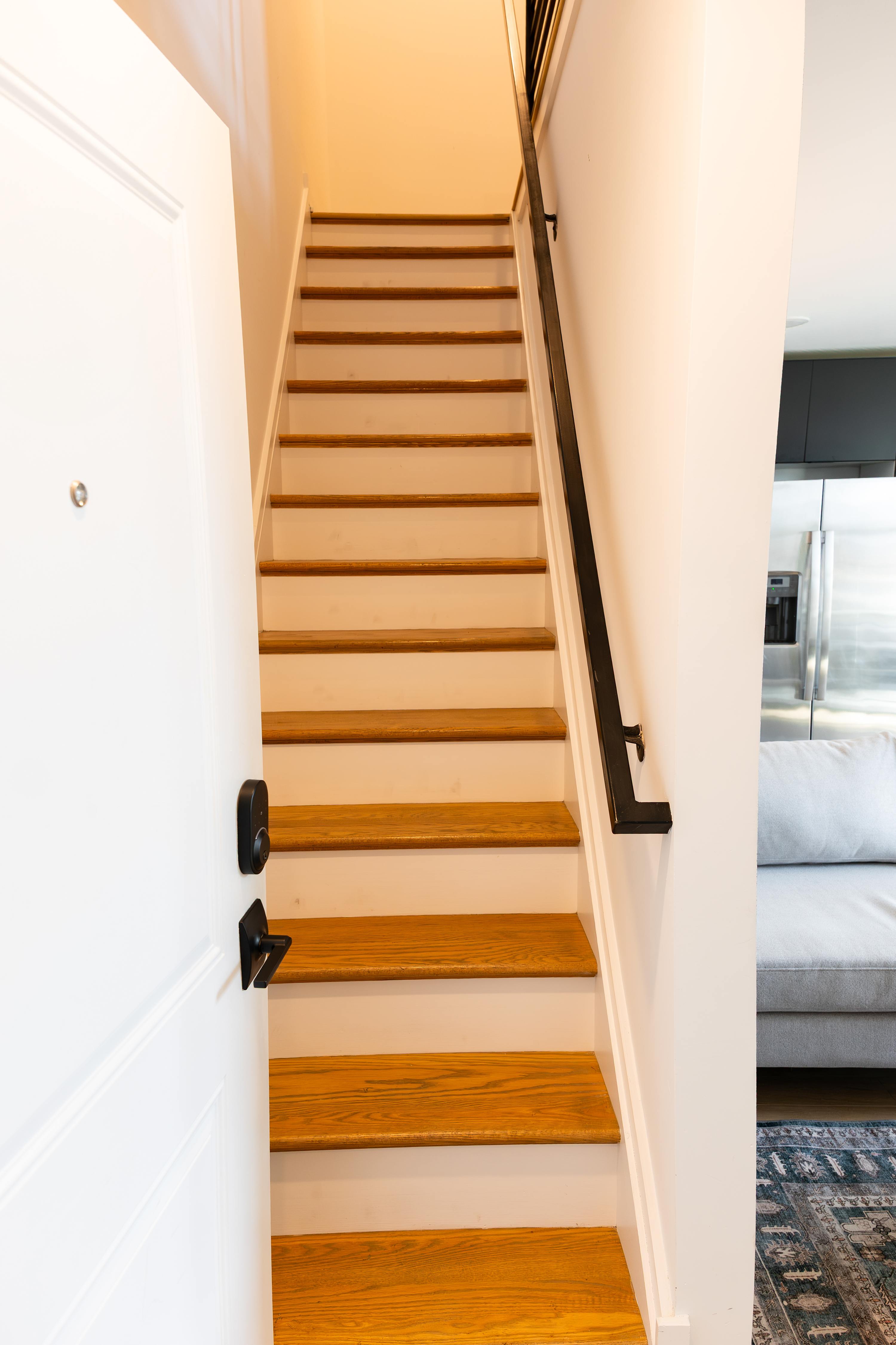 A welcoming foyer showcasing a wooden staircase with a black handrail, leading upstairs from an open white entrance door equipped with a smart lock.