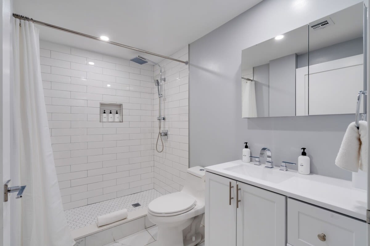 Bright bathroom featuring a walk-in shower with white subway tiles and a glass door, a white vanity with a single sink, and a mirrored medicine cabinet. The shower floor has white hexagonal tiles, contrasting with larger white floor tiles.