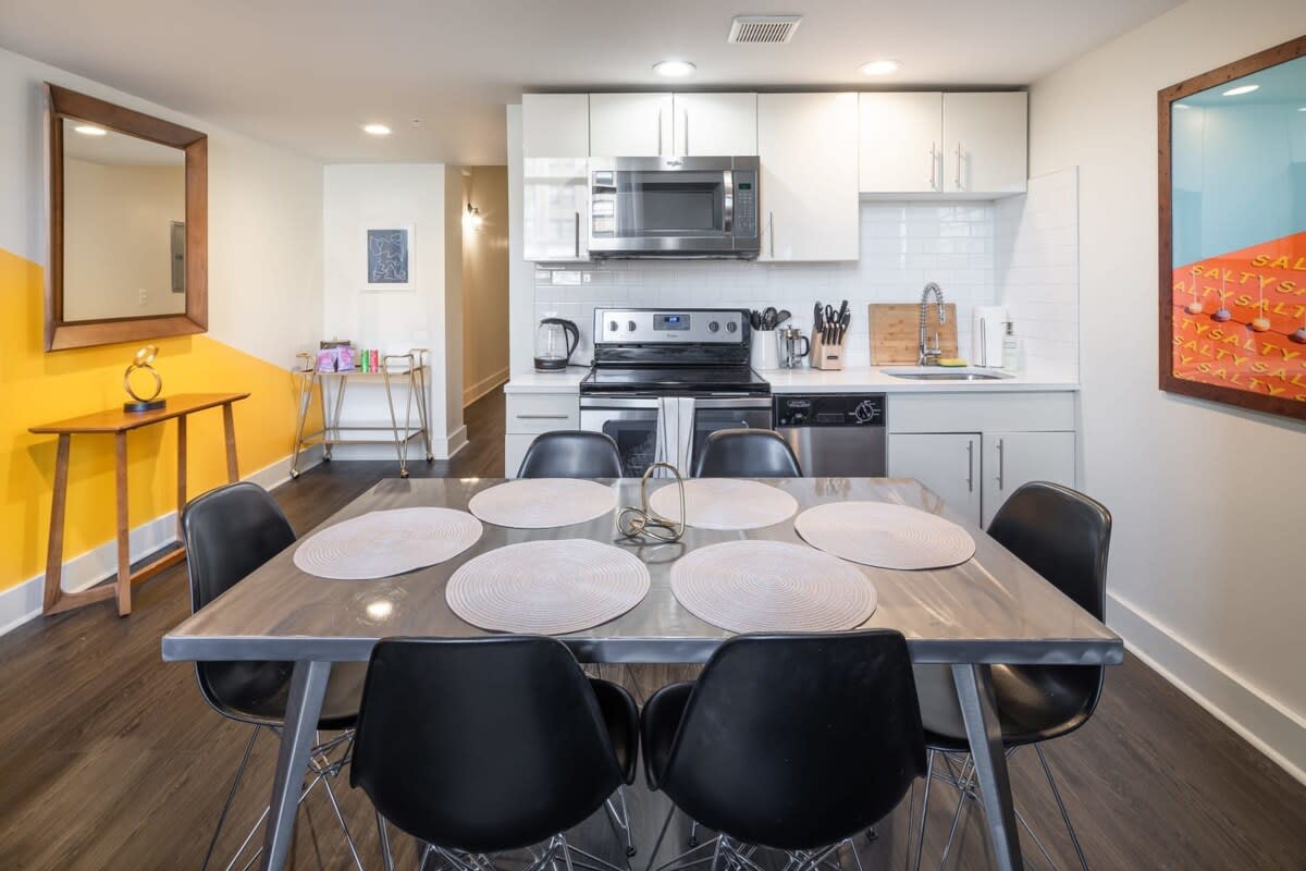 Modern kitchen with white cabinetry, subway tile backsplash, and stainless steel appliances, including a microwave, stove, and dishwasher. A large dining table with black chairs is centered in the foreground, with a yellow accent wall visible in the background.