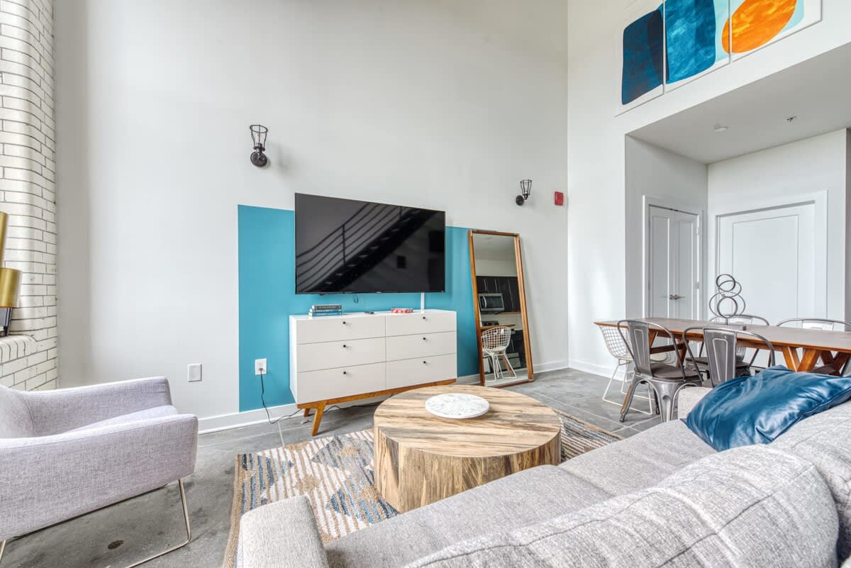 Spacious living area featuring a large flat-screen TV mounted above a white dresser against a teal accent wall, a light gray sofa, a round wooden coffee table, and a dining table with chairs in the background.