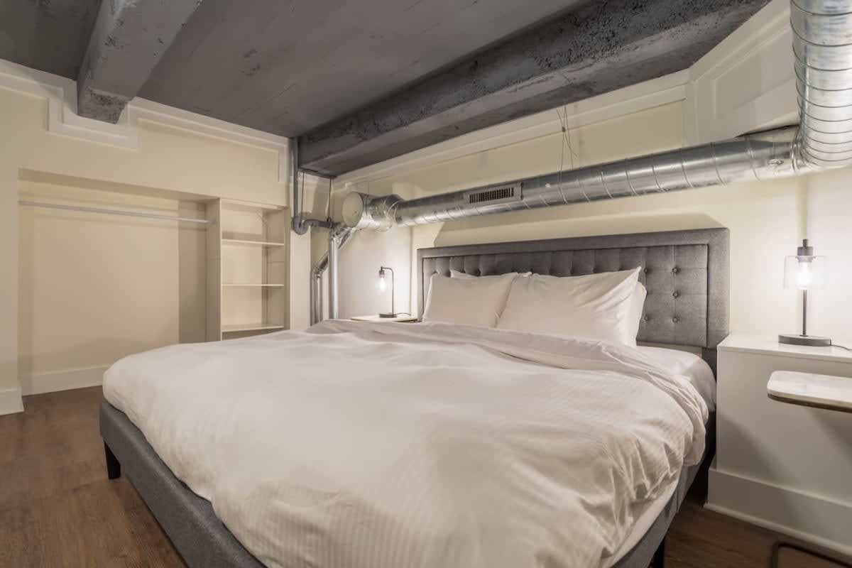 Close-up view of one of the gray upholstered queen beds in bedroom-1, featuring white bedding, dark wood nightstands with modern lamps, and visible industrial ductwork and ceiling beams.