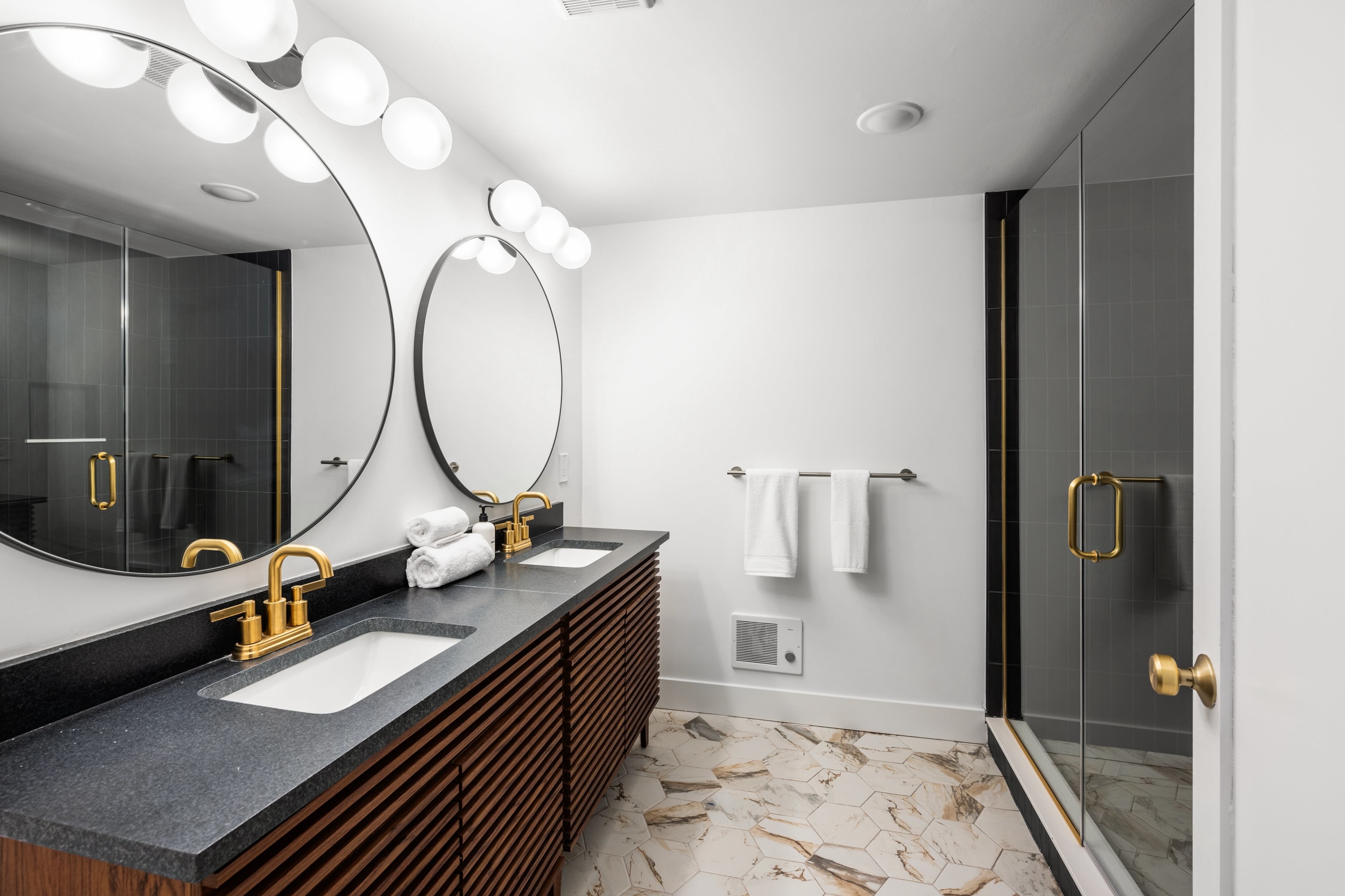 A contemporary bathroom with a double vanity featuring a dark countertop, gold faucets, and two round mirrors with globe lighting. A glass-enclosed shower with dark gray tiles and a gold handle is visible on the right.
