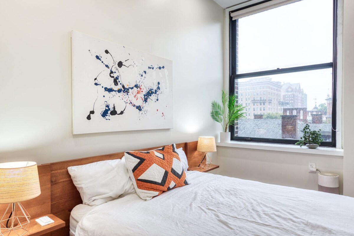 A cozy bedroom with a queen-sized bed featuring a wooden headboard, two floating nightstands with lamps, and a large abstract painting above the bed, with a window offering a city view.