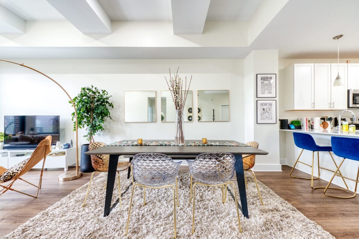 A contemporary dining area with a dark rectangular table, clear acrylic chairs with gold legs, and a shaggy beige rug, adjacent to a kitchen with white cabinets and blue bar stools.