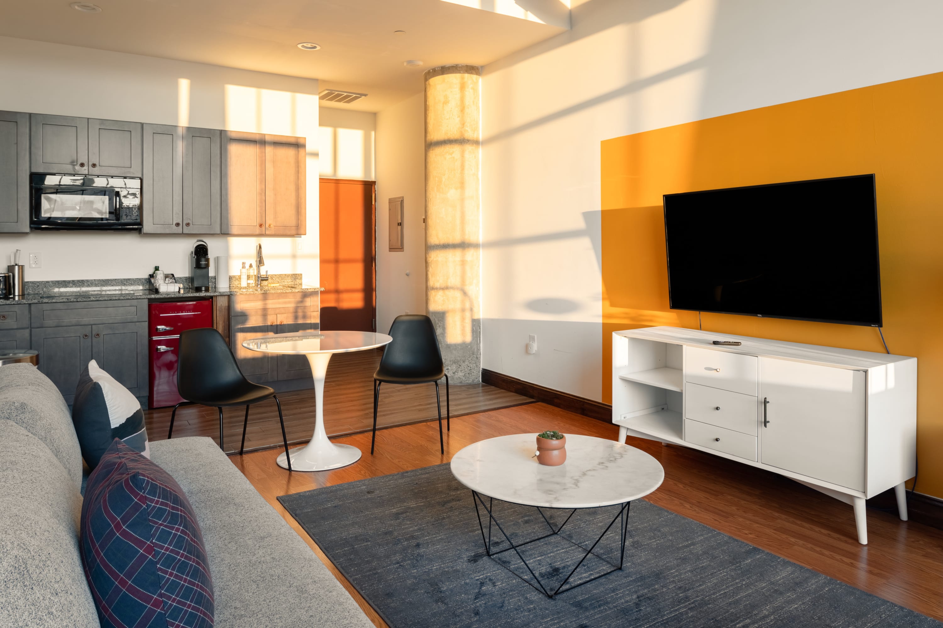 A cozy living area with a gray sofa and a white TV console with a large flat-screen TV against a wall featuring a bold yellow accent, adjacent to a compact kitchen with dark cabinets and a small dining table.
