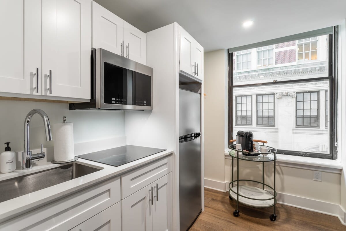 A well-lit kitchenette featuring white cabinets, a stainless steel microwave, and a two-burner electric cooktop integrated into the white countertop. A coffee station on a rolling cart is visible by the window, which has a room-darkening shade.