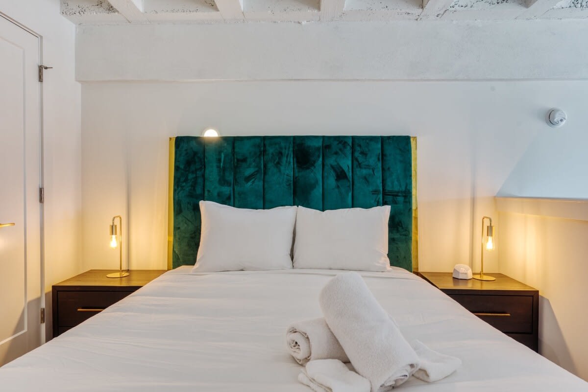 Cozy bedroom with a plush green velvet headboard and crisp white bedding, flanked by two dark wood nightstands with modern gold lamps. The room has white walls and a white exposed beam ceiling.