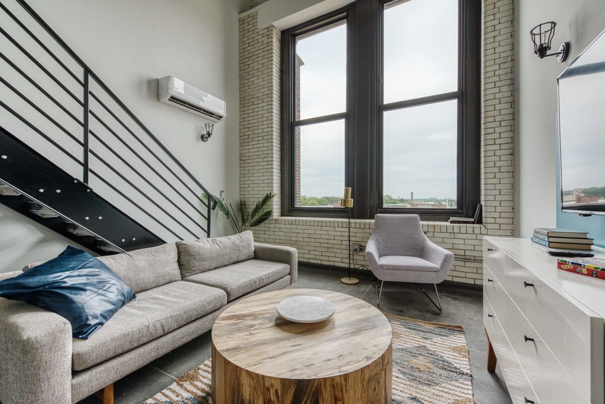 Contemporary living room with a light gray sofa, a round wooden coffee table, and a light gray armchair, all set on a patterned rug, with tall industrial windows and a metal staircase visible.