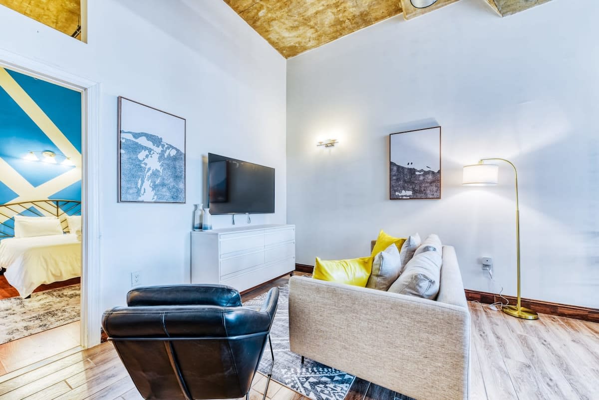 Living room showcasing a light gray sofa with yellow throw pillows, a black leather armchair, and a wall-mounted TV above a white console. A doorway leads to a bedroom with a blue accent wall.