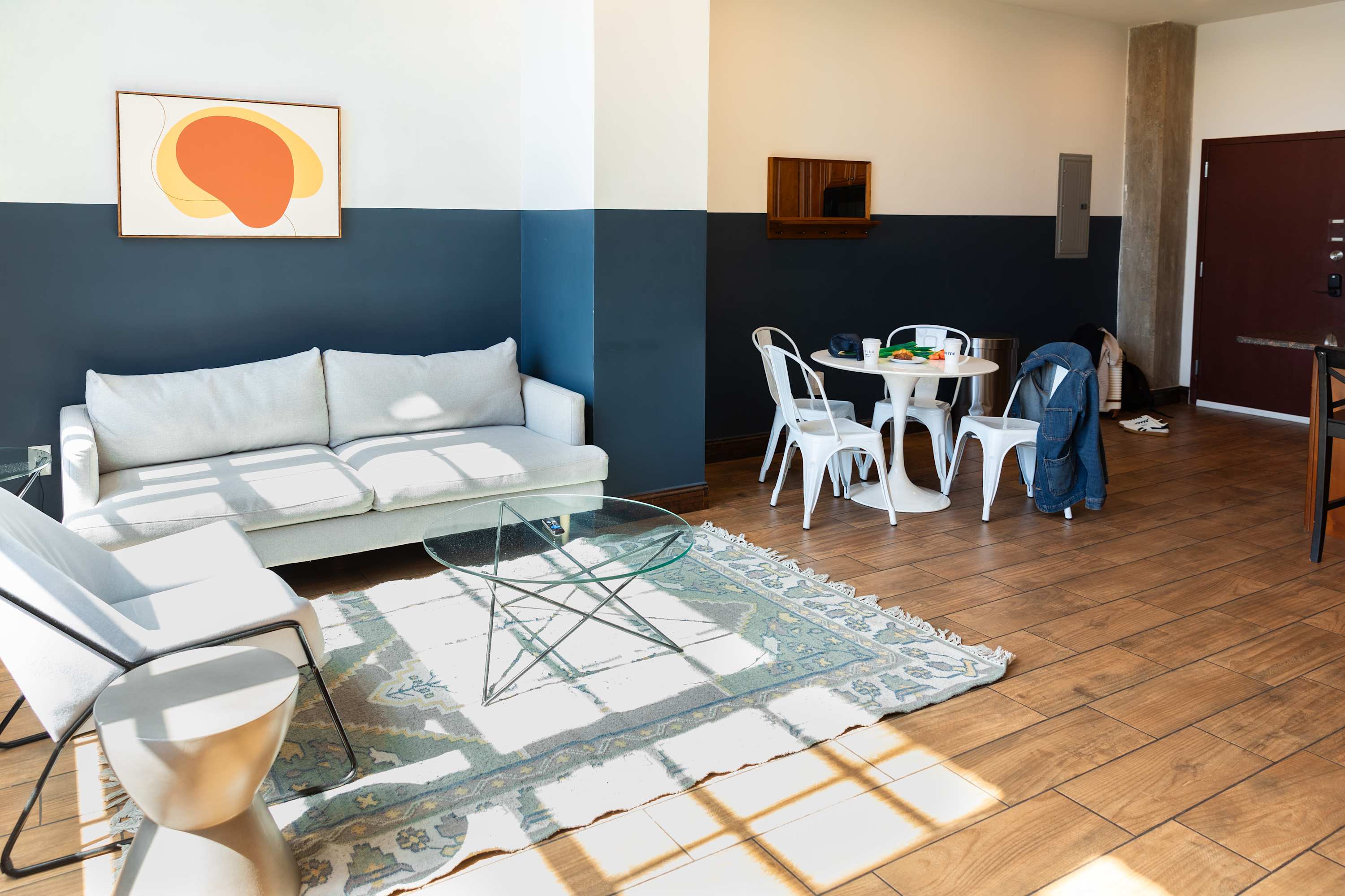 Open-concept living area with a light grey sofa, two armchairs, a glass coffee table over a patterned rug, and a dining area with a round white table and chairs, all set on hardwood floors against a two-tone wall.