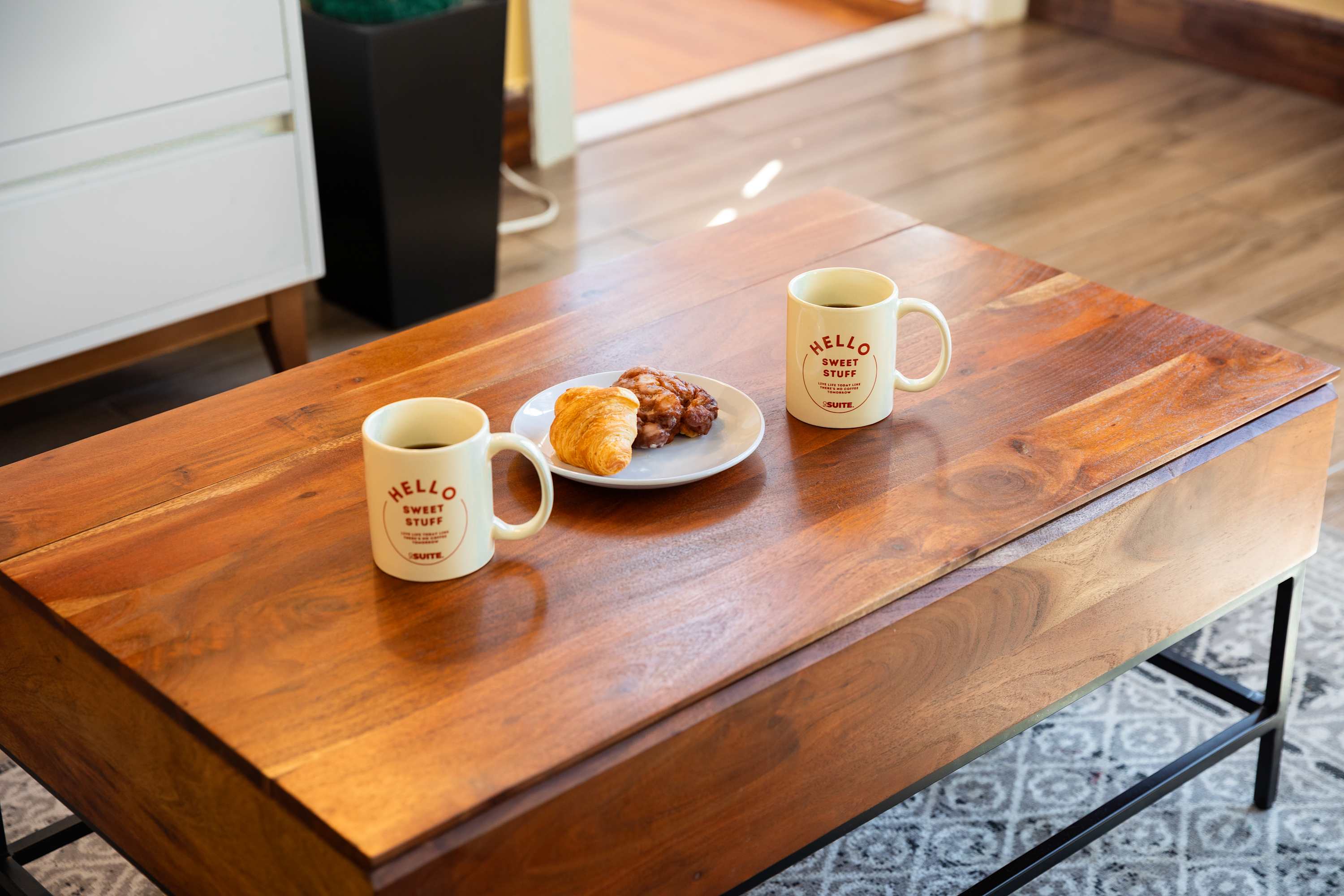 A close-up of a wooden coffee table in the living room, featuring two mugs with "HELLO SWEET STUFF" text and a plate with a croissant and pastry. The table sits on a patterned rug.