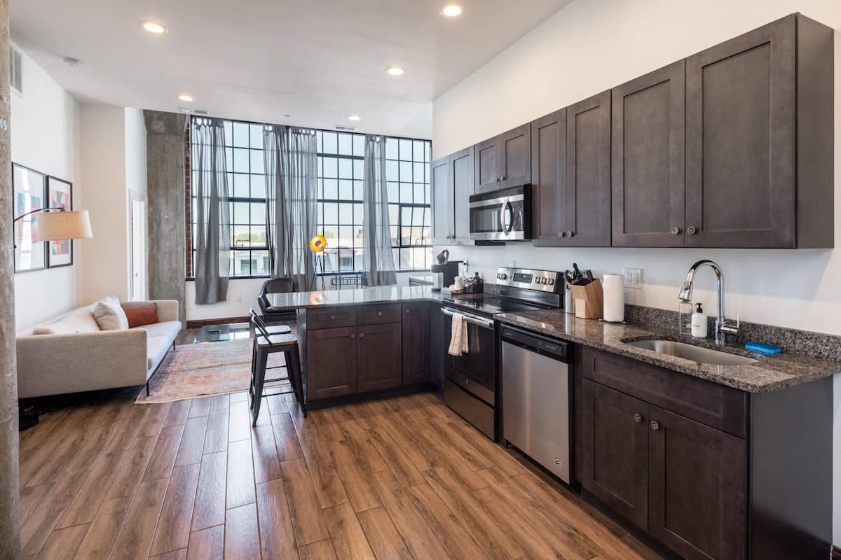 Modern kitchen with dark wood cabinets, granite countertops, and stainless steel appliances, open to a living area with large industrial-style windows and a sofa. The space features warm hardwood floors.