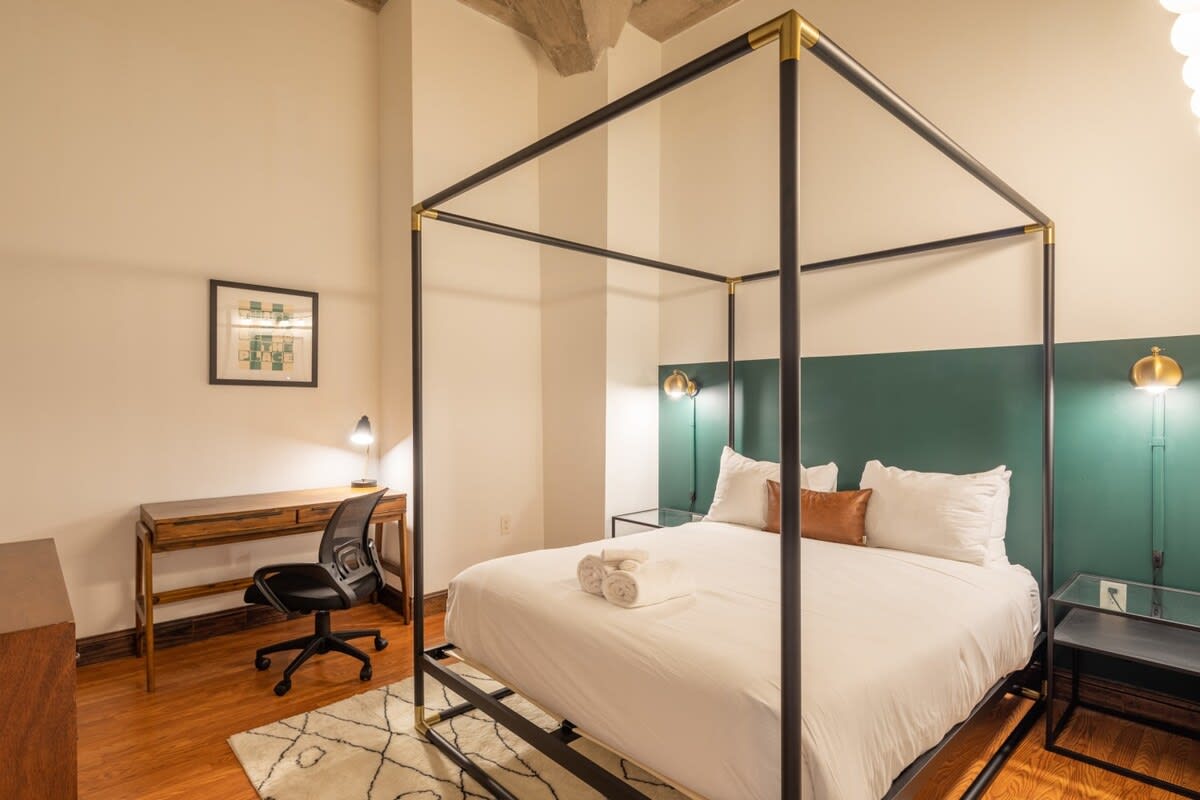 Elegant bedroom featuring a black and gold canopy bed with white bedding, a dark green accent wall behind the headboard, and a wooden desk with an office chair, all set on warm hardwood floors.