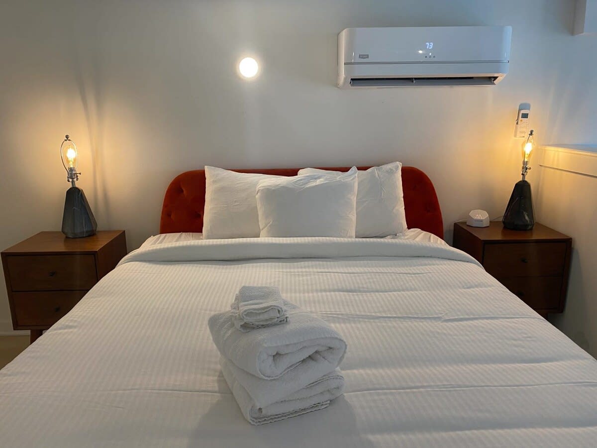 Cozy bedroom featuring a red velvet headboard on a queen-sized bed with white linens, flanked by two wooden nightstands with industrial-style lamps. A wall-mounted AC unit is above the bed.