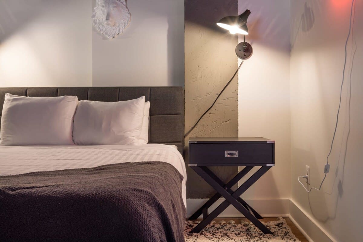 Cozy bedroom with a grey upholstered headboard and white and grey bedding. A black nightstand with an X-shaped base and a wall-mounted lamp are positioned next to the bed, with a textured rug underneath.
