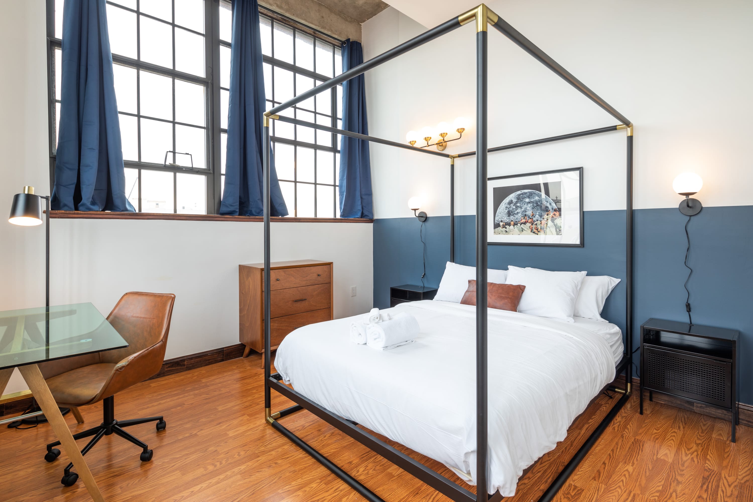 Stylish bedroom with a black metal four-poster bed, white bedding, and a two-tone wall featuring a dark blue lower half. Large industrial windows with blue curtains are visible, and a desk with a glass top and a brown office chair is in the corner.