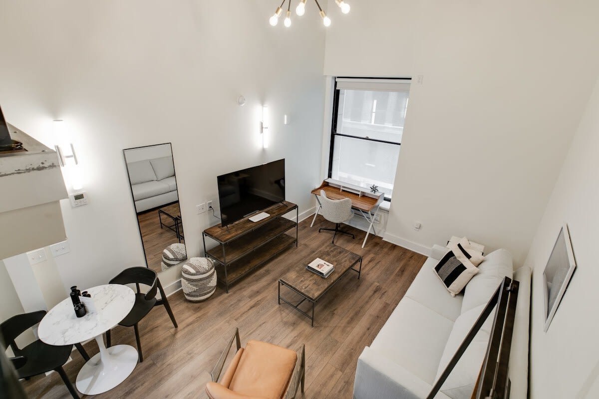 Overhead view of a bright living area featuring a white sofa, a brown leather armchair, a coffee table, and a dining table with two chairs. A desk and chair are positioned by a large window, and a flat-screen TV is on a wooden media console.