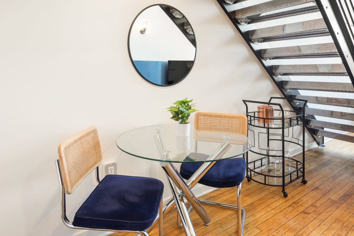 A compact dining area featuring a round glass-top table with chrome legs and two chairs with blue velvet seats and cane backs, positioned next to a black metal staircase and a bar cart.