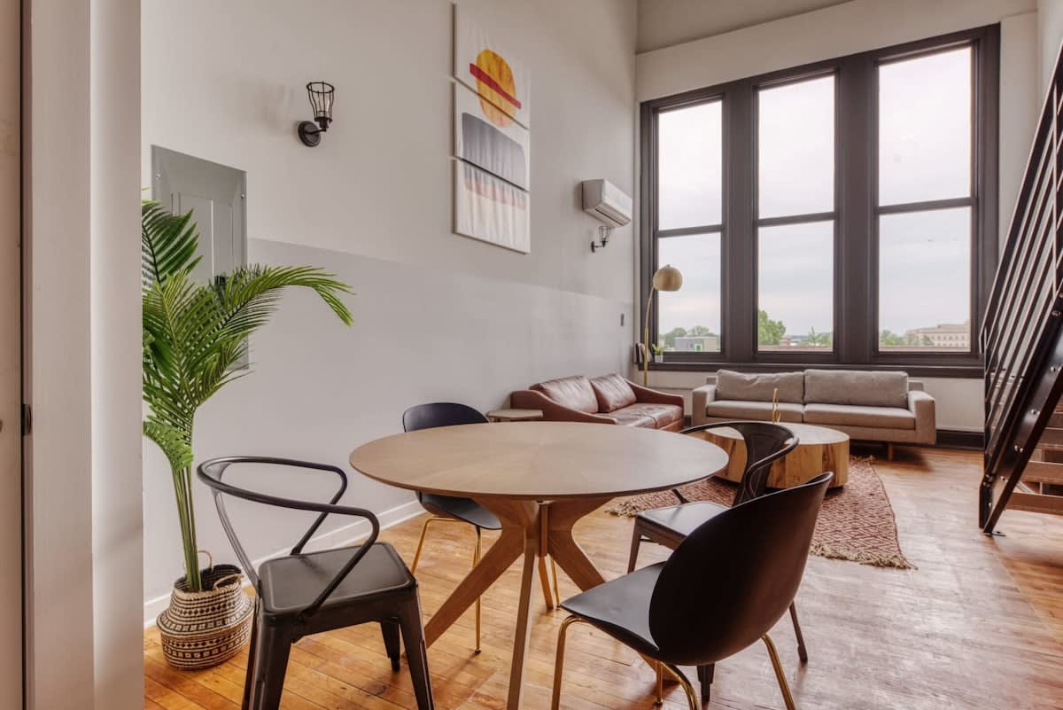 Open-concept living and dining area featuring a round wooden dining table with black chairs, a brown leather sofa, a grey sofa, and a round coffee table, with large windows overlooking the city.