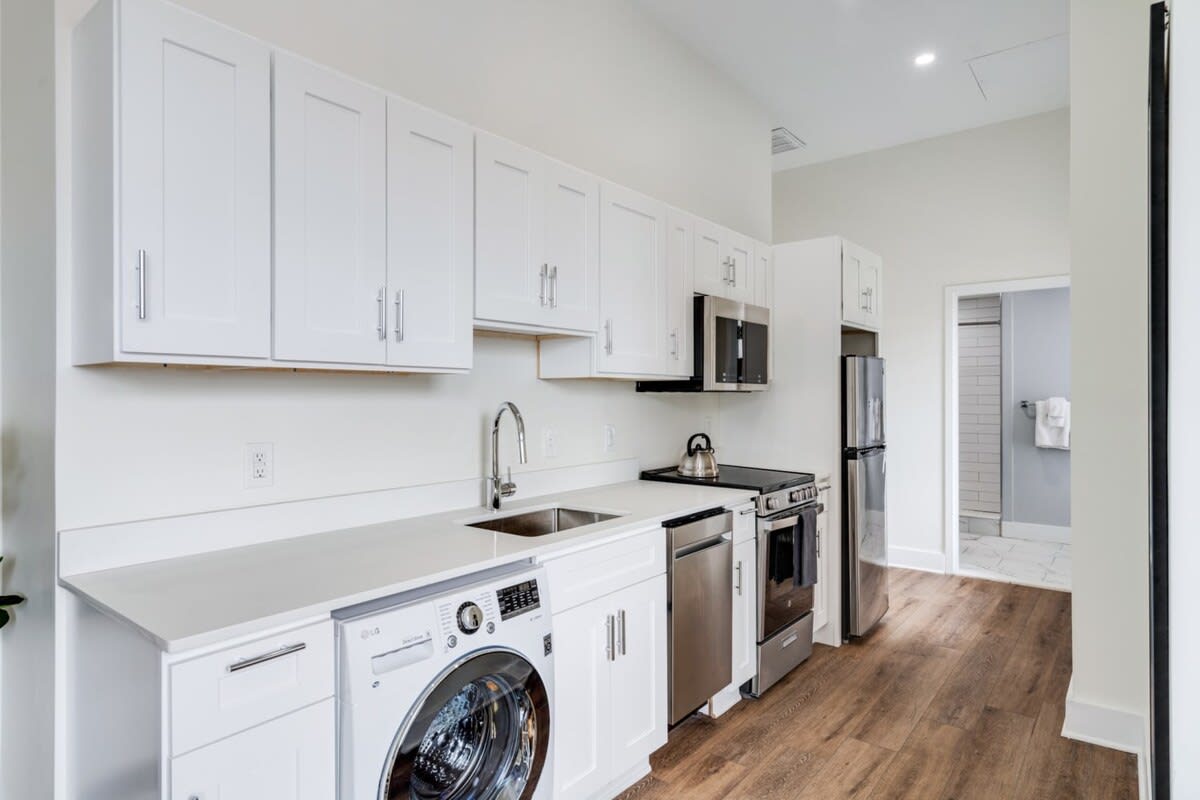 Modern kitchen with white shaker cabinets, white countertops, stainless steel appliances including a refrigerator, microwave, and range, and an in-unit washer/dryer.