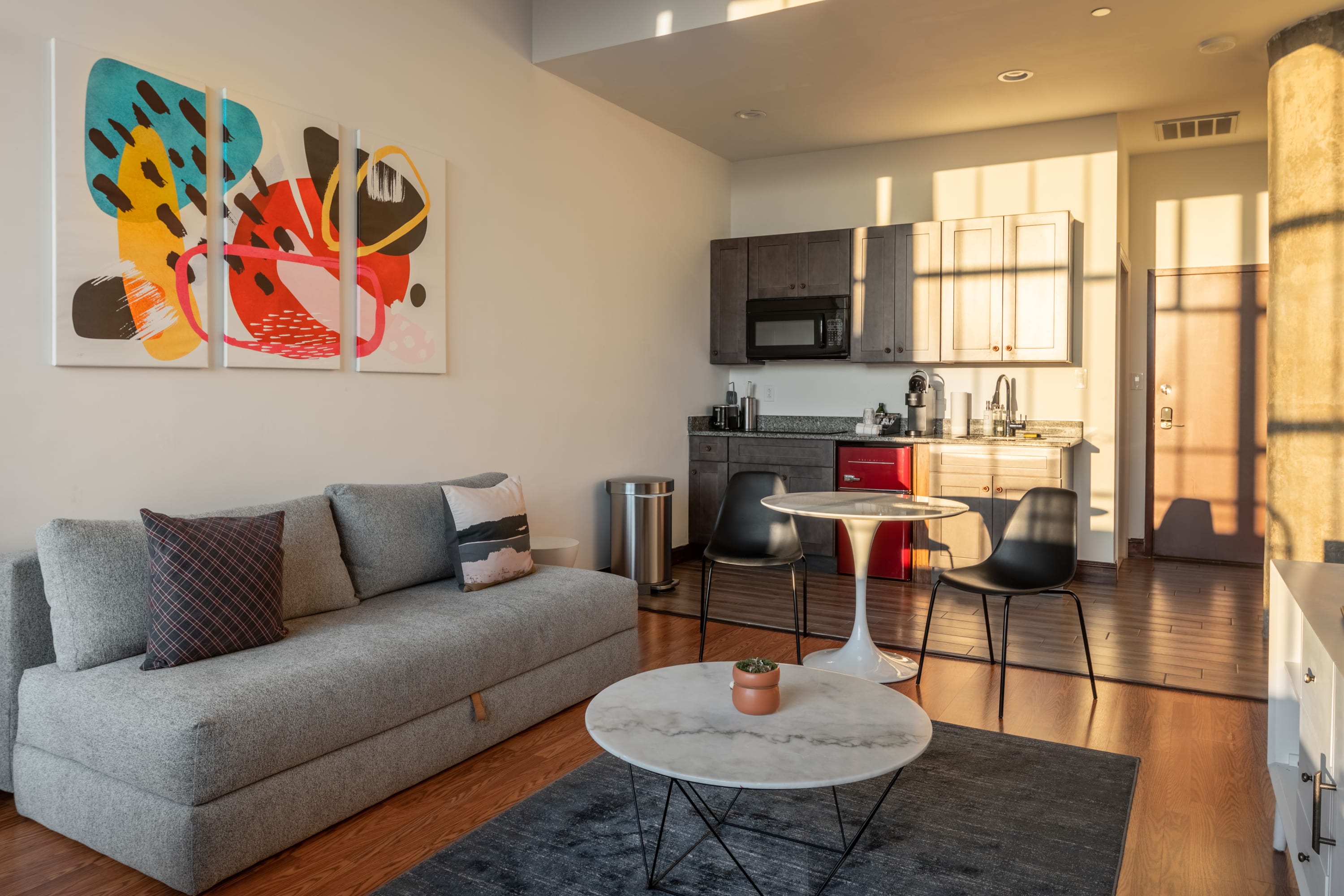 A modern living space featuring a gray sofa with colorful throw pillows, a white marble-top coffee table, and a compact kitchen with dark gray cabinets, granite countertops, and a vibrant red mini-fridge.