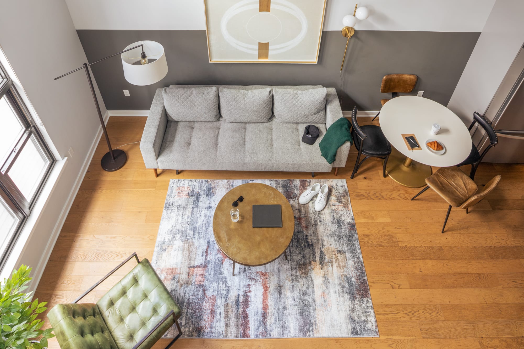 An aerial view of the open-concept living and dining area, showcasing a light gray sofa, a round wooden coffee table on an abstract rug, and a small white dining table with mixed chairs. The space features hardwood floors and a two-tone wall.